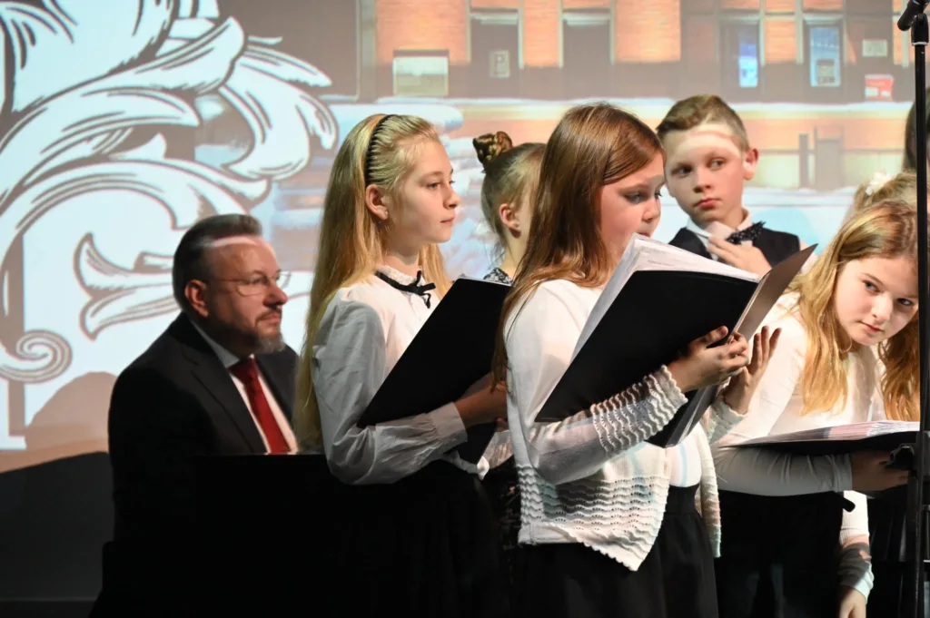 Na zdjęciu dzieci i młodzież z chóru Most the Music z Rudy Śląskiej w trakcie występu. Ubrani na galowo, w rękach trzymają zeszyty z nutami. Po lewej stronie chórmistrz.