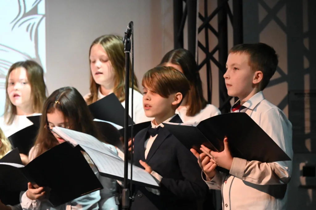 Na zdjęciu dzieci i młodzież z chóru Most the Music z Rudy Śląskiej w trakcie występu. Ubrani na galowo, w rękach trzymają zeszyty z nutami.