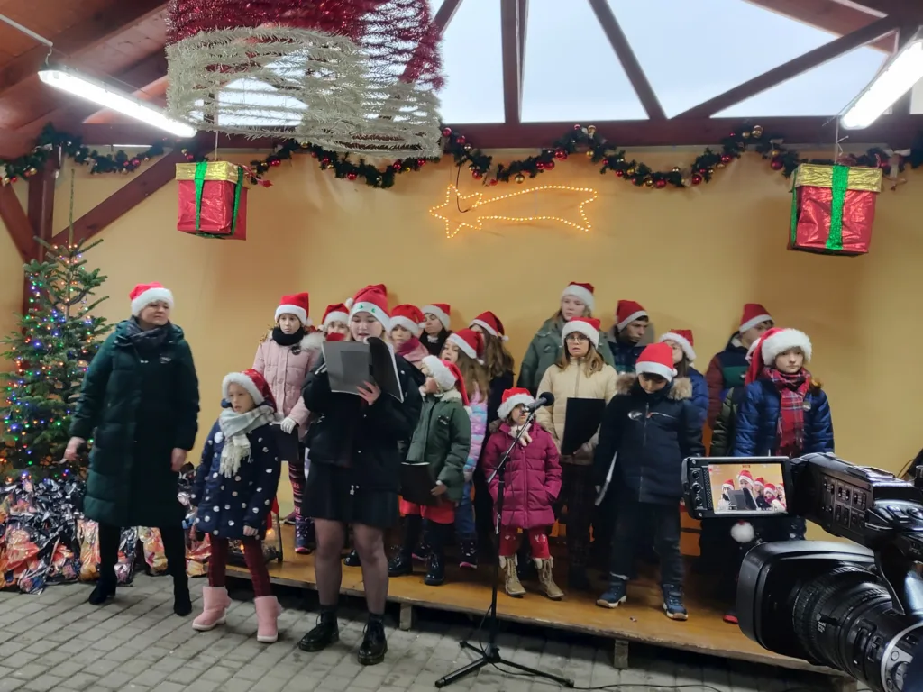 Chór Most the Music z Dziewkowic na scenie na zewnątrz budynku podczas jarmarku świątecznego. Grupa dzieci i młodzieży oraz chórmistrzyni w czasie śpiewu. Wszyscy mają na głowach czapki Mikołaja.
