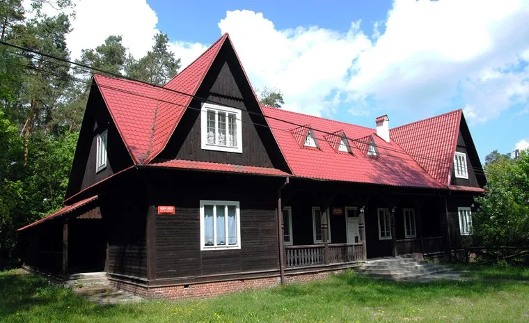 Drewniany, ciemny budynek z czerwonym dachem, w otoczeniu zieleni.