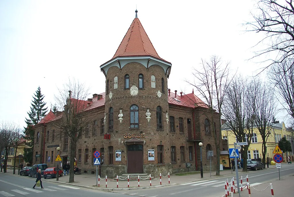 Zdjęcie przedstawia dwukondygnacyjny budynek z cegły, stojący u zbiegu ulic. Jego elementem charakterystycznym jest wieżyczka z czerwonym dachem.