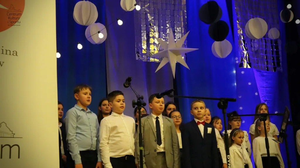 Na scenie ubrane na galowo dzieci z chóru Most the Music w trakcie występu. W tle granatowa dekoracja z połyskującymi srebrnymi elementami (wiszące kółka, gwiazdy) i lampkami.