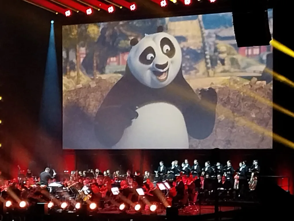 Zdjęcie uroczyście oświetlonej sceny. Artyści w trakcie występu. W tle kadr z filmu animowanego "Kung Fu Panda".