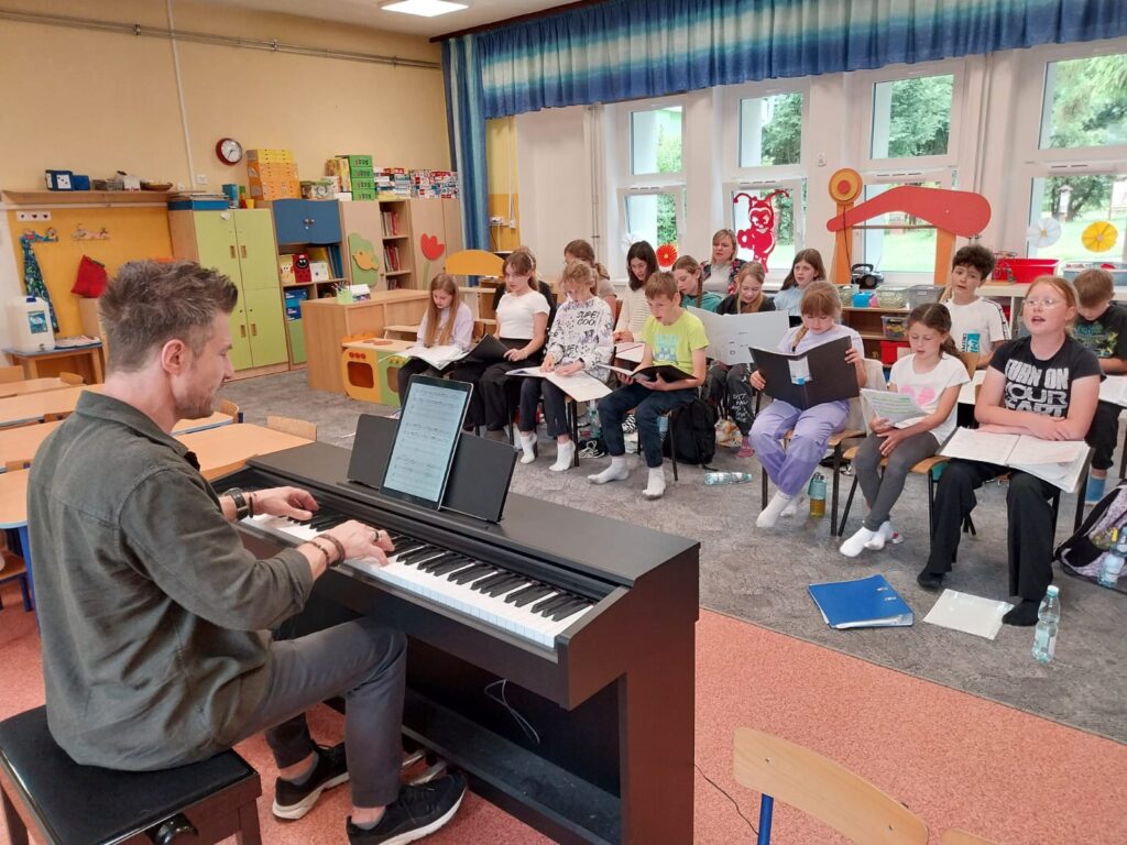 Dzieci i młodzież podczas warsztatów śpiewu. Adam Sztaba przy instrumencie klawiszowym.