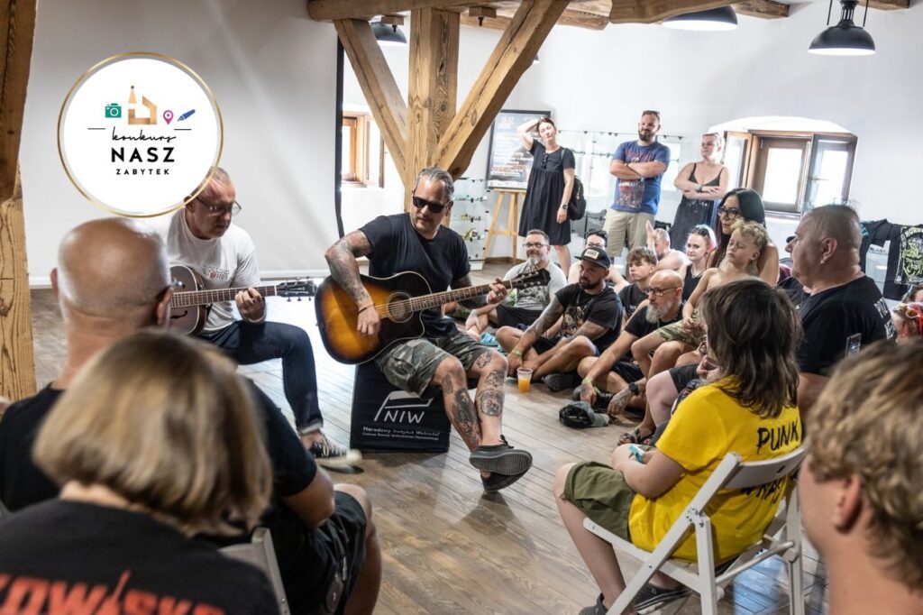 zrewaloryzowany spichlerz wewnątrz. Dwóch mężczyzn gra na gitarze. Wokół siedzą i stoją widzowie.